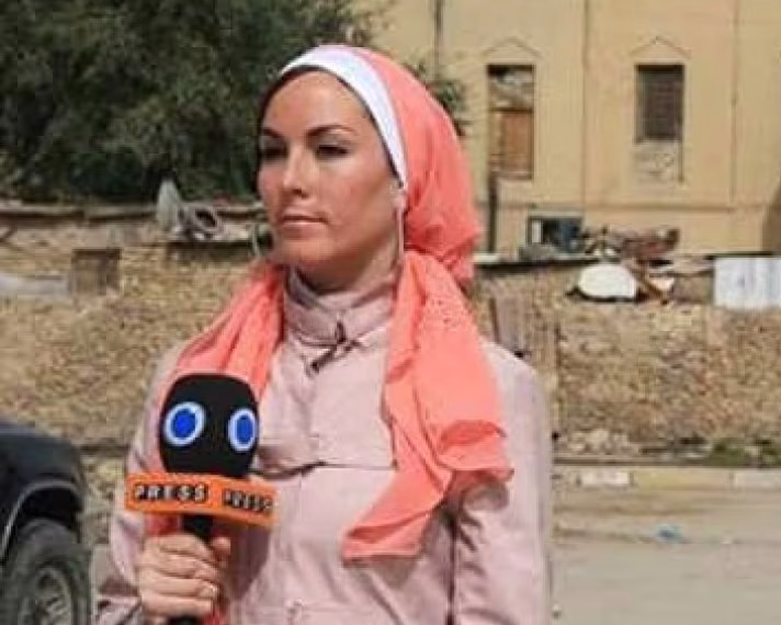 Amanda Lindhout wearing a pink outfit and orange headscarf, holding a Press TV microphone while reporting outdoors in Baghdad, Iraq.